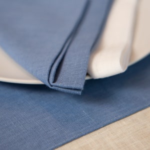 May include: A close-up of a table setting featuring a folded blue linen napkin, a white plate, and a rolled white napkin. The blue napkin is on a blue linen placemat, with a beige linen surface in the background. The setting suggests a dining theme.