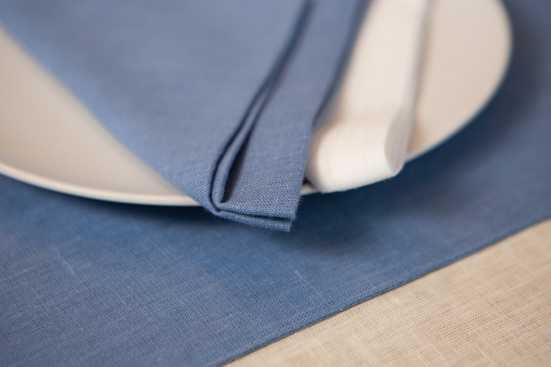 May include: A close-up of a table setting featuring a folded blue linen napkin, a white plate, and a rolled white napkin. The blue napkin is on a blue linen placemat, with a beige linen surface in the background. The setting suggests a dining theme.