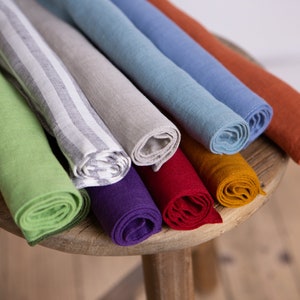 May include: A collection of rolled linen napkins in various colors, including green, gray, blue, and orange, displayed on a wooden stool. Some napkins have striped patterns.