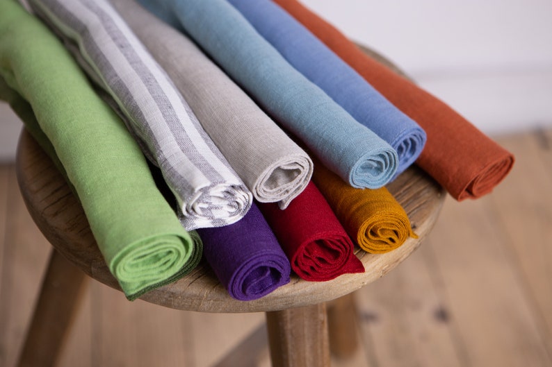 May include: A collection of rolled linen napkins in various colors, including green, gray, blue, and orange, displayed on a wooden stool. Some napkins have striped patterns.