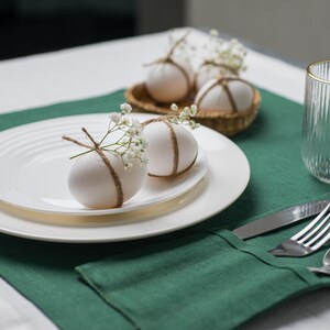 May include: A table setting with white plates, a green placemat, and a clear glass with a gold rim. Decorated eggs with twine and flowers sit on the plates and in a small basket. Silverware is neatly arranged on a green napkin.