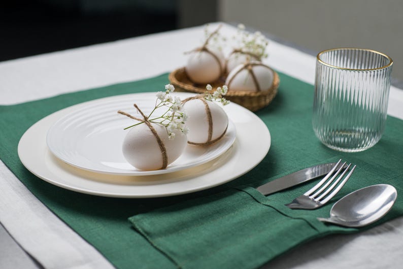 May include: A table setting with white plates, a green placemat, and a clear glass with a gold rim. Decorated eggs with twine and flowers sit on the plates and in a small basket. Silverware is neatly arranged on a green napkin.