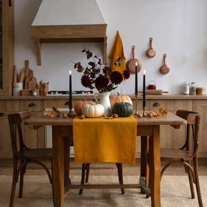 May include: A wooden dining table features a mustard yellow linen runner, a white ceramic pitcher filled with dark red dahlias, three pumpkins in shades of white, peach, and green, and two black taper candles in brass holders with scattered nuts.