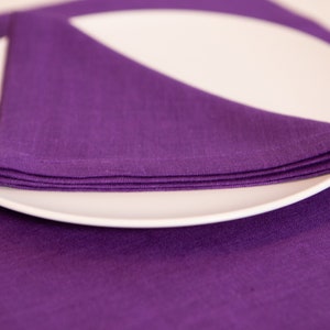 May include: A white, oval-shaped plate with a folded purple linen napkin. The napkin is neatly arranged on the plate, with the corner of the napkin pointing upwards. The plate rests on a matching purple placemat.