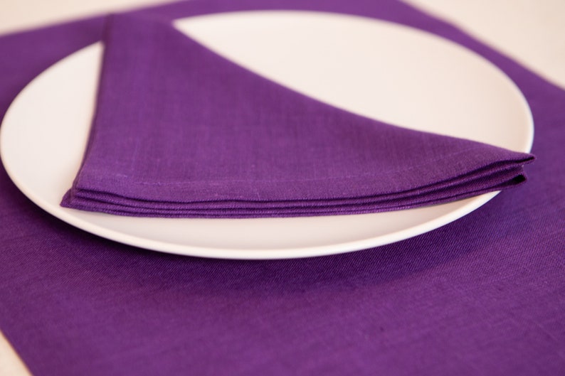 May include: A white, oval-shaped plate with a folded purple linen napkin. The napkin is neatly arranged on the plate, with the corner of the napkin pointing upwards. The plate rests on a matching purple placemat.