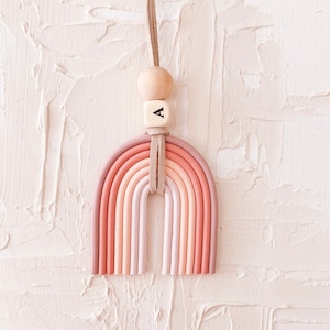 Könnte beinhalten: Ein rosafarbenes und pfirsichfarbenes regenbogenförmiges Tonornament mit einer Holzperle und einem Buchstaben "A" Anhänger, der an einer braunen Schnur hängt.