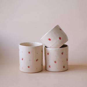 Peut inclure: Trois tasses en céramique blanc cassé avec un motif répété de petites fraises rouges et de feuilles vertes. Les tasses sont cylindriques et empilées, l'une étant inclinée sur le côté. Le fond est de couleur rose clair uni.