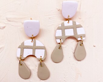 May include: A pair of statement earrings with a geometric design. The earrings are made of white and beige clay and feature a checkered pattern. The earrings are suspended from a small, white clay disc.