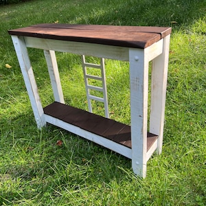 May include: White console table with a brown top and a ladder-like design on the front. The table is sitting on a green grassy lawn.