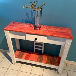 May include: A rustic console table with a red-stained wooden top and a white-painted base. The table features a drawer with a black knob and a lower shelf. A glass vase with dried plants sits on top. The table measures approximately 48 inches long.