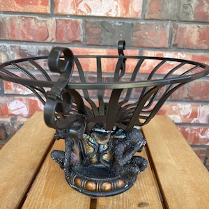 May include: A decorative black metal fruit basket with a detailed base featuring monkey figures. The basket has a wide, open design with curved handles and sits on a wooden surface.