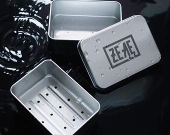 May include: Three silver metal soap dishes. One dish has a drainage design with holes. Another dish has a lid with the word "ZEAE" printed on it. The dishes are on a black surface.