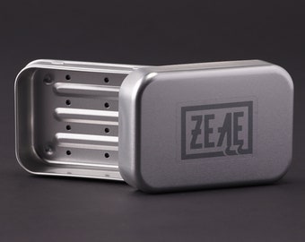 May include: A silver metal soap dish with a raised, ridged design and drainage holes. The lid has the word "ZEAL" printed on it.
