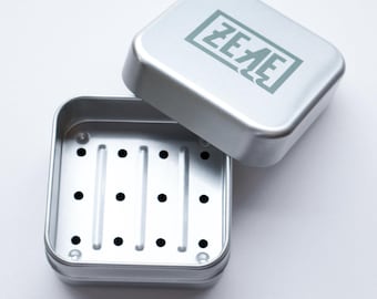May include: A silver soap dish with a raised, square design and a drain hole. The lid has the word "ZEZE" printed on it.