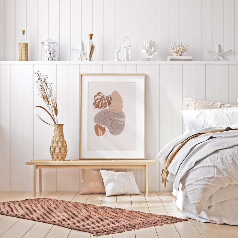 May include: A framed print with a minimalist abstract design featuring brown and beige leaves and shapes on a light pink background. The print is hanging on a white wall above a wooden bench with a brown and beige rug in front of it.