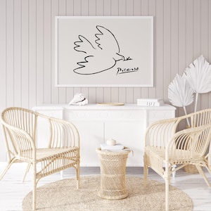 May include: A black and white line drawing of a dove in flight with the signature "Picasso" below. The artwork is framed and hanging on a white wall above a white dresser with two wicker chairs and a wicker side table.