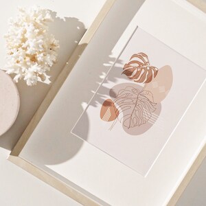 May include: A framed print with a minimalist design featuring a brown line drawing of a tropical leaf with overlapping abstract shapes in shades of brown and beige.