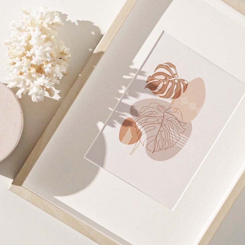May include: A framed print with a minimalist design featuring a brown line drawing of a tropical leaf with overlapping abstract shapes in shades of brown and beige.