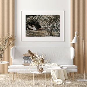 May include: A white couch with a stack of pillows and a vase of dried flowers on a white coffee table. A white lamp is in the corner of the room. A framed print of a painting of a garden with white flowers and two people sitting on chairs is on the wall behind the couch.
