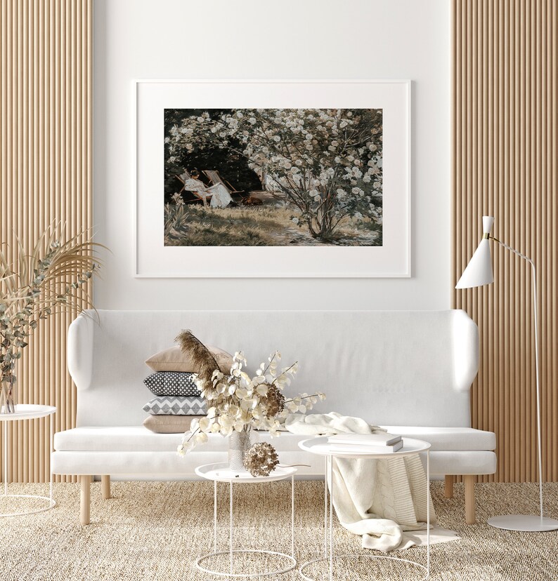 May include: A white couch with a stack of pillows and a vase of dried flowers on a white coffee table. A white lamp is in the corner of the room. A framed print of a painting of a garden with white flowers and two people sitting on chairs is on the wall behind the couch.