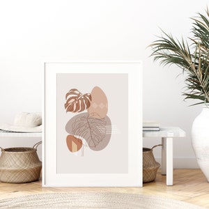 May include: A minimalist art print featuring a brown and beige abstract design with leaves and geometric shapes. The print is framed in a white frame and is hanging on a white wall.