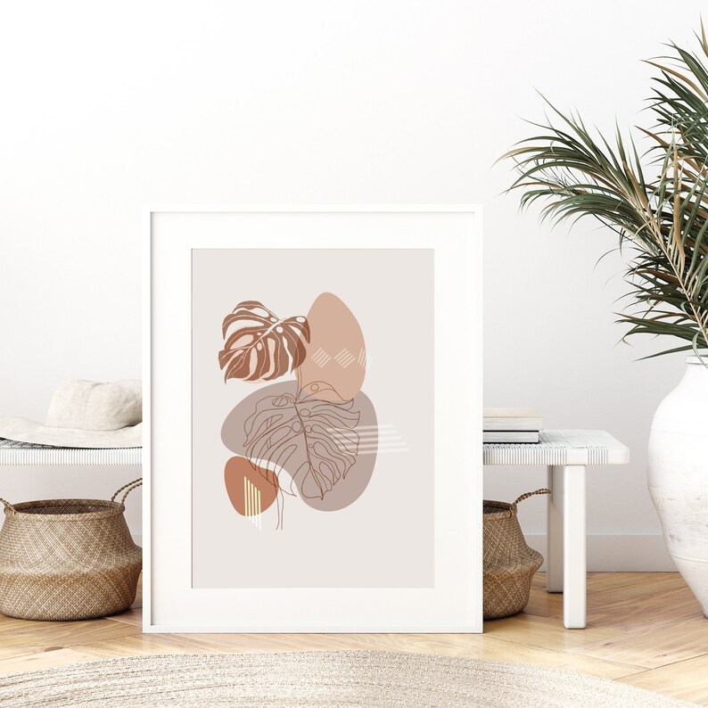May include: A minimalist art print featuring a brown and beige abstract design with leaves and geometric shapes. The print is framed in a white frame and is hanging on a white wall.