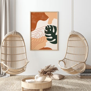 May include: Two hanging wicker chairs with a framed abstract print featuring a green leaf and orange and brown shapes. The print is hanging on a white wall.