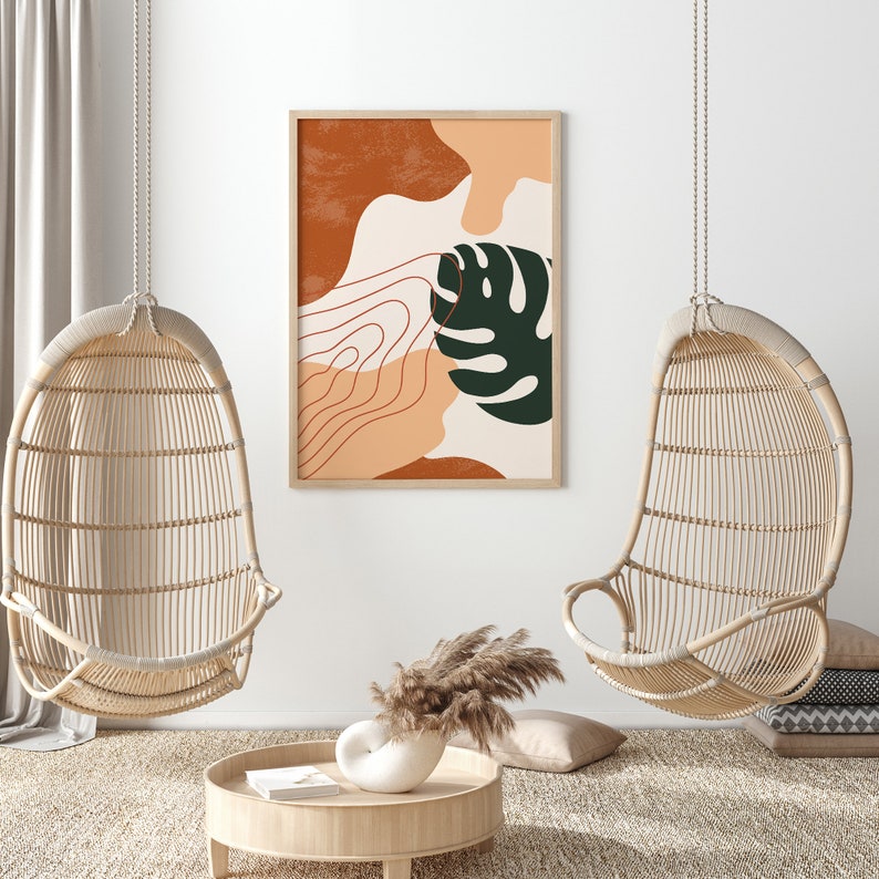 May include: Two hanging wicker chairs with a framed abstract print featuring a green leaf and orange and brown shapes. The print is hanging on a white wall.