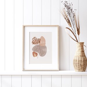 May include: A framed print with a minimalist design featuring brown and beige abstract shapes and line drawings of leaves. The print is displayed on a white shelf with a vase of dried flowers.