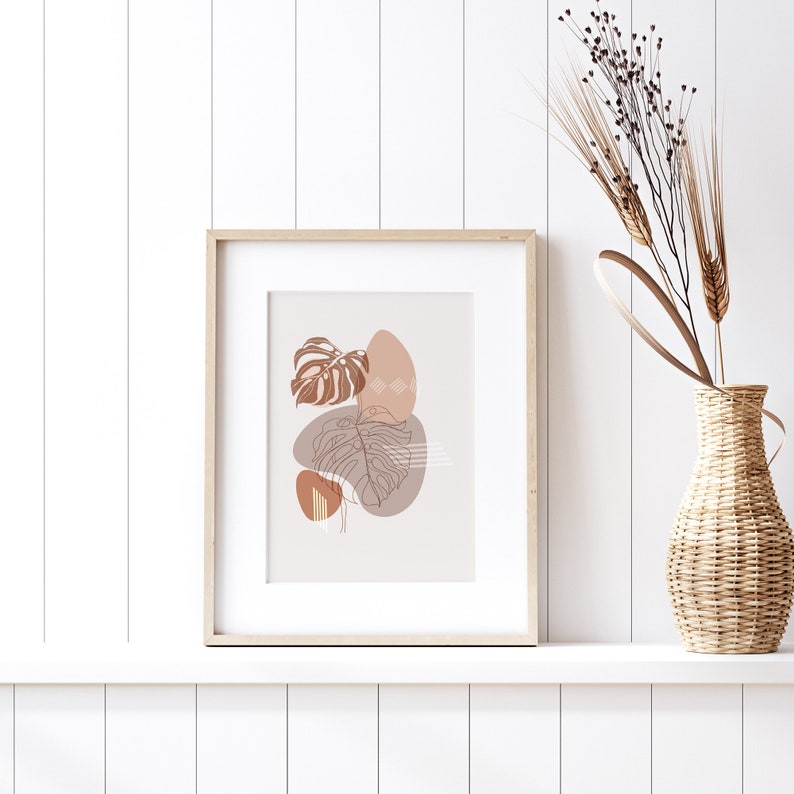 May include: A framed print with a minimalist design featuring brown and beige abstract shapes and line drawings of leaves. The print is displayed on a white shelf with a vase of dried flowers.