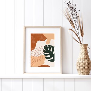 May include: A framed print with an abstract design featuring a green leaf, brown and orange shapes, and white lines. The print is in a light wood frame and is hanging on a white wall.