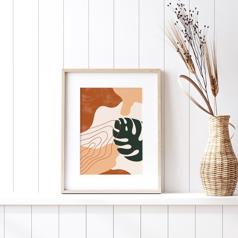 May include: A framed print with an abstract design featuring a green leaf, brown and orange shapes, and white lines. The print is in a light wood frame and is hanging on a white wall.