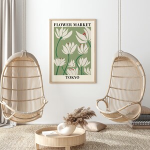 Puede incluir: Una impresi&oacute;n floral verde y blanca con el texto "FLOWER MARKET TOKYO" en un marco de madera. La impresi&oacute;n est&aacute; colgada en una pared blanca entre dos sillas de mimbre colgantes.