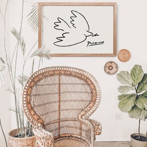 May include: A framed black and white line drawing of a dove with the signature "Picasso" below it. The frame is brown wood and the drawing is on a white background. The frame is hanging on a white wall above a wicker chair.