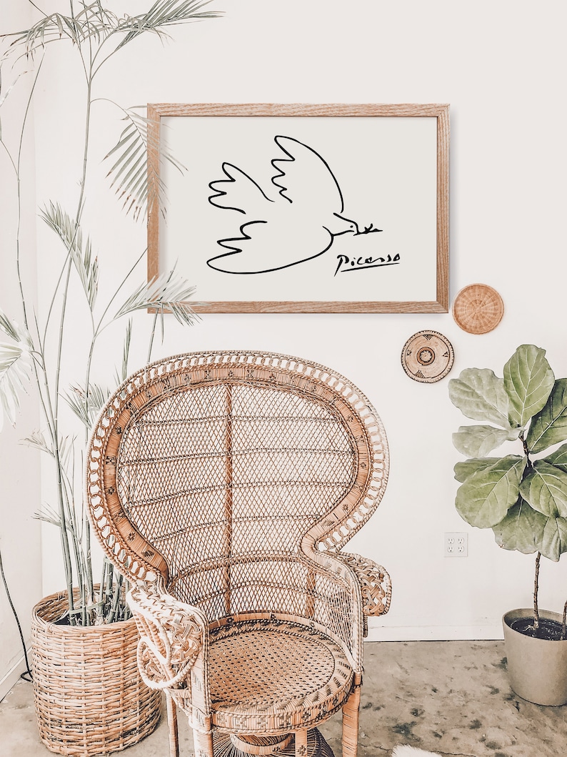 May include: A framed black and white line drawing of a dove with the signature "Picasso" below it. The frame is brown wood and the drawing is on a white background. The frame is hanging on a white wall above a wicker chair.