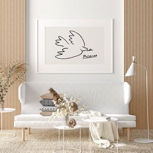 May include: A white sofa with a stack of pillows and a white coffee table with a white blanket on it. A framed black and white line drawing of a dove by Picasso hangs on the wall above the sofa.