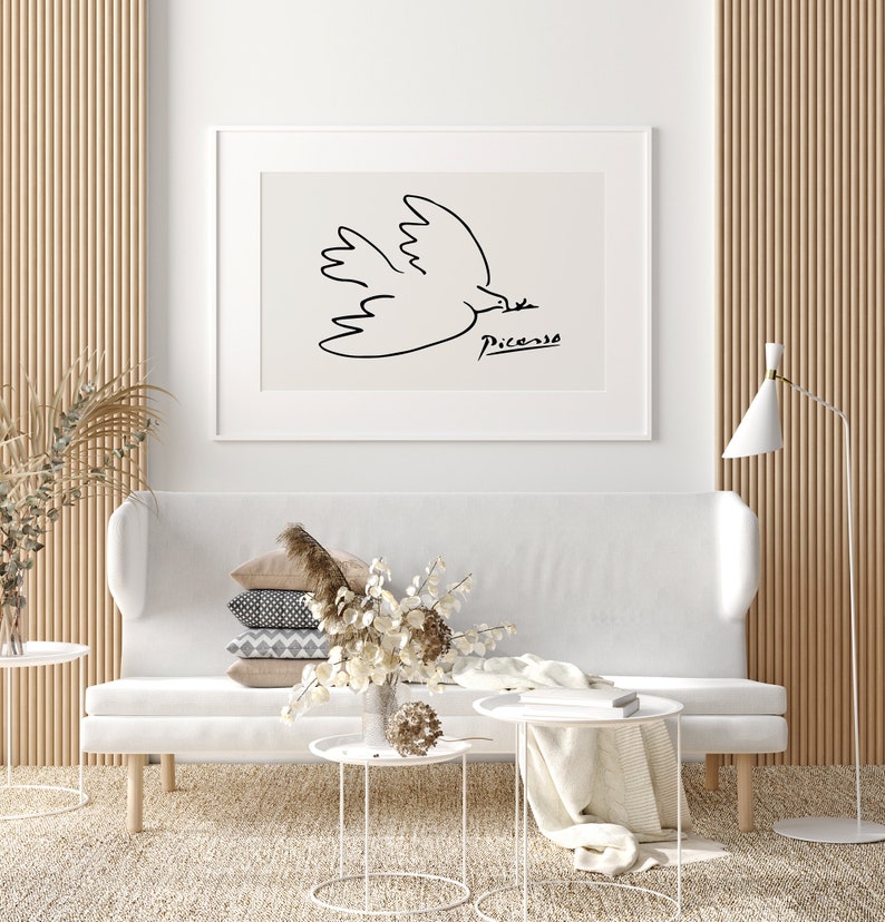 May include: A white sofa with a stack of pillows and a white coffee table with a white blanket on it. A framed black and white line drawing of a dove by Picasso hangs on the wall above the sofa.