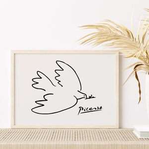 May include: A black and white line drawing of a dove in flight, with the artist's signature "Picasso" below. The drawing is framed in a light wood frame and displayed on a white wall.