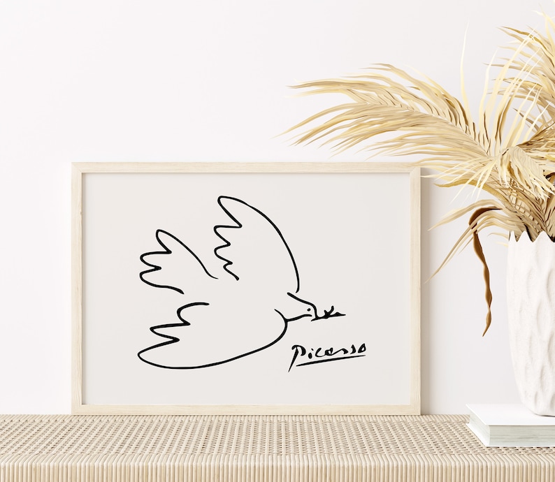 May include: A black and white line drawing of a dove in flight, with the artist's signature "Picasso" below. The drawing is framed in a light wood frame and displayed on a white wall.