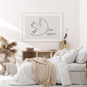 May include: A white bed with a beige throw blanket and white pillows. A framed print of a black and white line drawing of a dove by Picasso hangs above the bed.