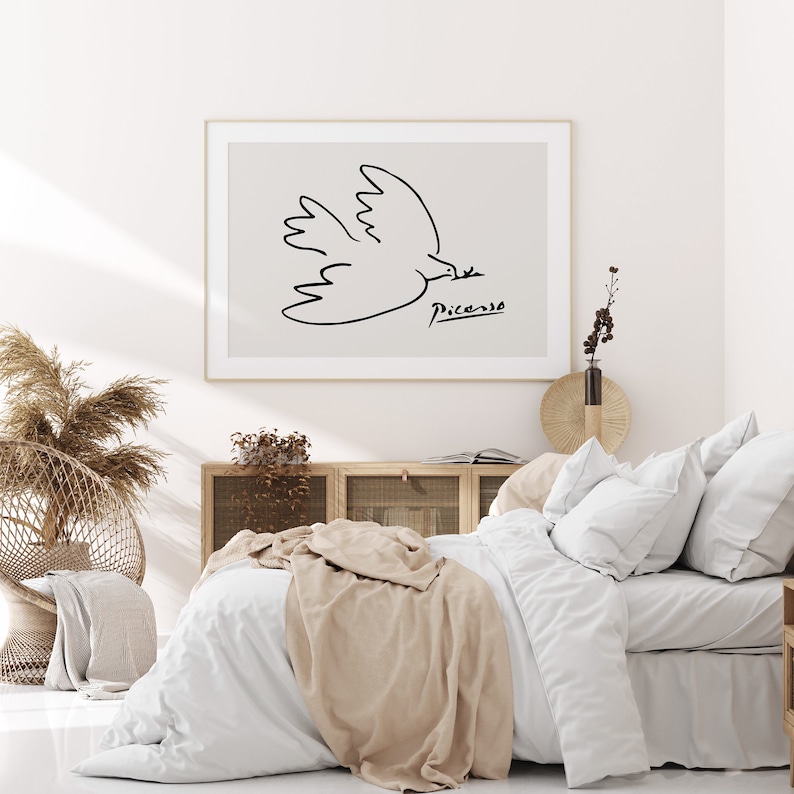 May include: A white bed with a beige throw blanket and white pillows. A framed print of a black and white line drawing of a dove by Picasso hangs above the bed.