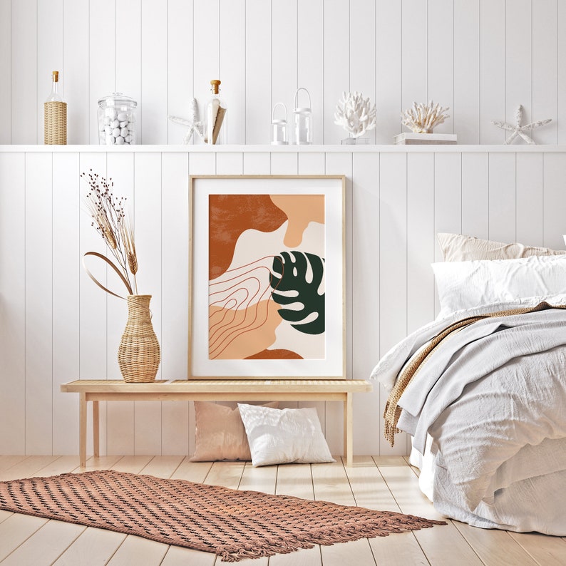 May include: A framed abstract print with a brown, orange, and green color scheme. The print features a stylized leaf and organic shapes. The print is hanging on a white wall above a wooden bench and a brown rug.