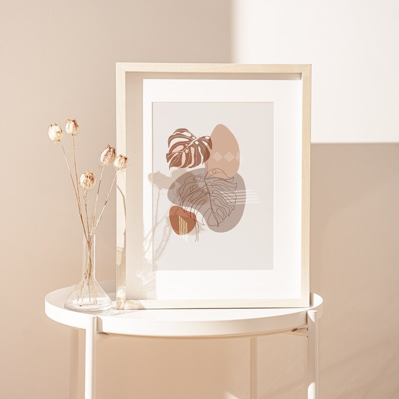 May include: A framed print with a minimalist design featuring abstract shapes and line drawings of leaves in shades of brown and beige. The print is displayed on a white side table with a vase of dried flowers.