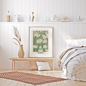 Puede incluir: Un p&oacute;ster enmarcado con fondo verde y flores blancas estilizadas. El p&oacute;ster dice "FLOWER MARKET TOKYO". El p&oacute;ster est&aacute; colgado en una pared blanca sobre un banco de madera y una alfombra marr&oacute;n.