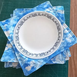 May include: A stack of blue and white quilted placemats with a white plate with a blue and white floral border on top.