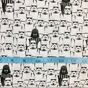 Peut inclure: Un tissu blanc avec un motif répété de casques de Stormtrooper noirs et blancs de la franchise Star Wars. Un casque de Dark Vador noir et blanc est également inclus dans le motif. Un ruban à mesurer bleu se trouve en bas de l'image.