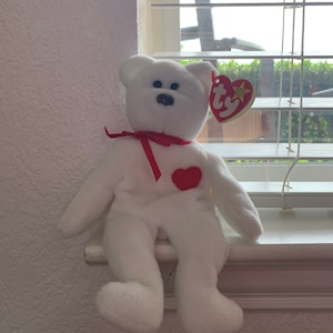 May include: White plush teddy bear with a red heart on its chest and a red ribbon around its neck. The bear is sitting on a windowsill. The bear has a Ty tag attached to its ear.
