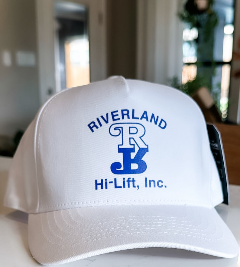 Puede incluir: Gorra de b&eacute;isbol blanca con un logotipo azul que dice "Riverland R Hi-Lift, Inc."