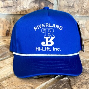Puede incluir: Una gorra de b&eacute;isbol azul real con ribete de cuerda blanca. La gorra tiene un logotipo bordado en blanco que dice "Riverland" con una "R" y una "B" estilizadas dentro de un c&iacute;rculo. Debajo del logotipo, dice "Hi-Lift, Inc."