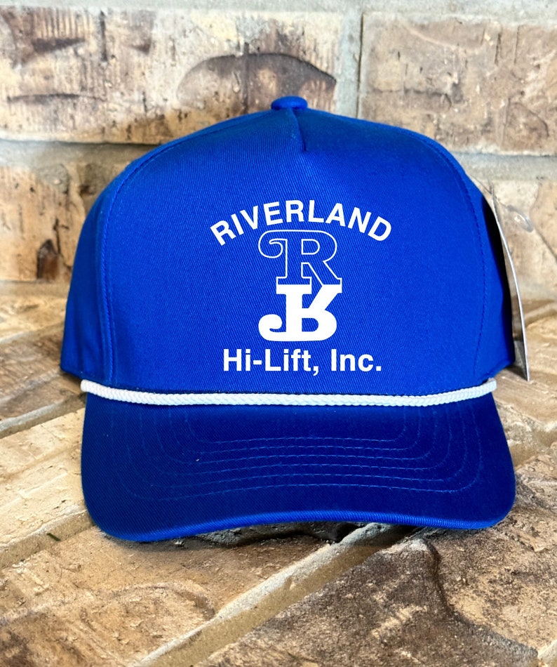 Puede incluir: Una gorra de b&eacute;isbol azul real con ribete de cuerda blanca. La gorra tiene un logotipo bordado en blanco que dice "Riverland" con una "R" y una "B" estilizadas dentro de un c&iacute;rculo. Debajo del logotipo, dice "Hi-Lift, Inc."